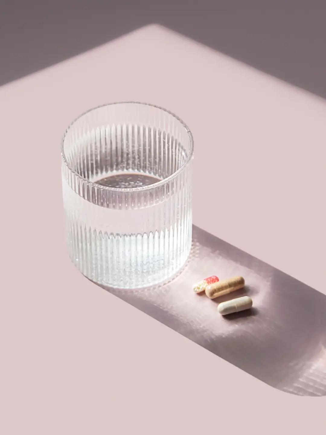 Stabilità dei probiotici: capsule e umidità | Encanto Nutraceutica Capsule di integratore accanto a un bicchiere d’acqua, in luce naturale su fondo rosa