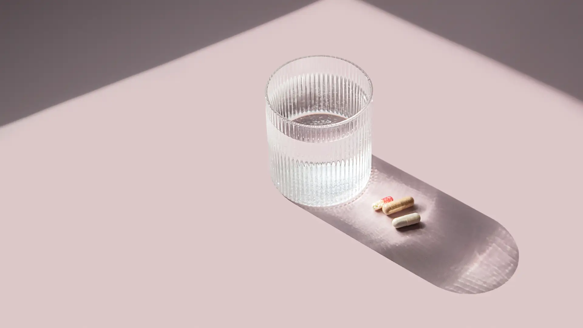 Capsule di integratore accanto a un bicchiere d’acqua, in luce naturale su fondo rosa